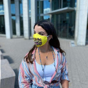 May include: A woman wearing a yellow face mask with a colorful floral pattern, a striped shirt, and blue jeans. She is wearing a beaded necklace with a peace sign charm.