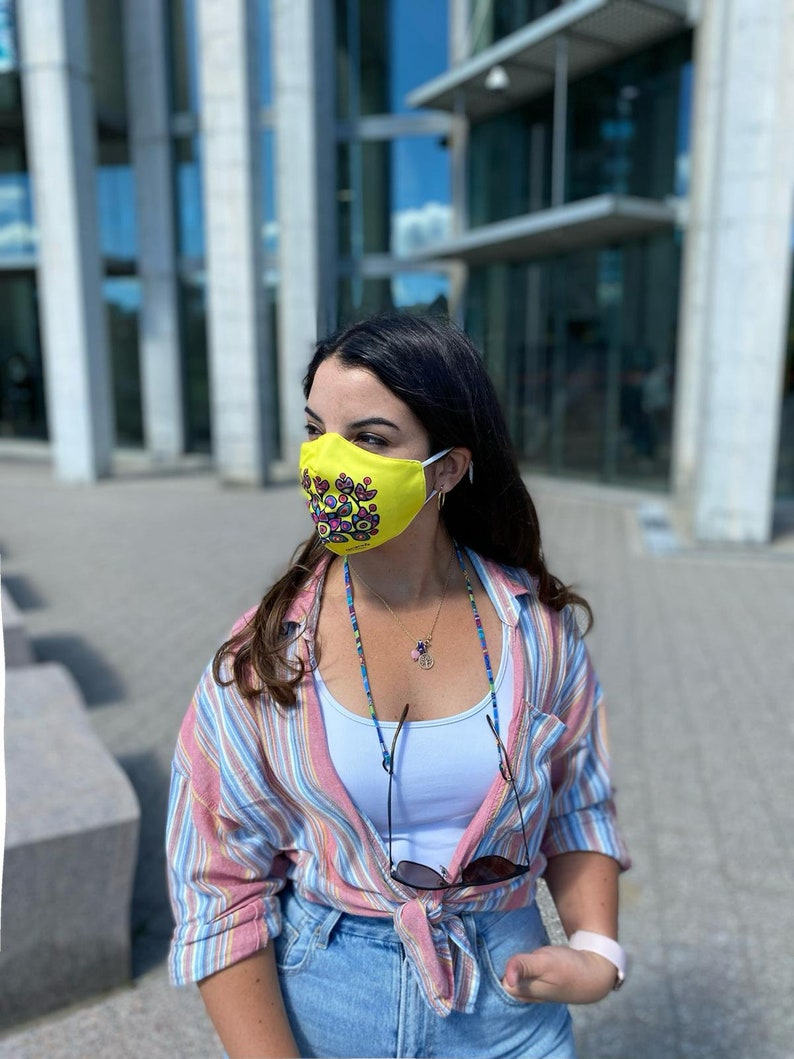May include: A woman wearing a yellow face mask with a colorful floral pattern, a striped shirt, and blue jeans. She is wearing a beaded necklace with a peace sign charm.