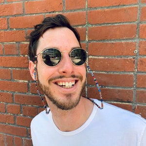 May include: A man with a beard is smiling and wearing round, dark sunglasses with a colorful patterned strap. He is wearing a white t-shirt and standing in front of a brick wall.