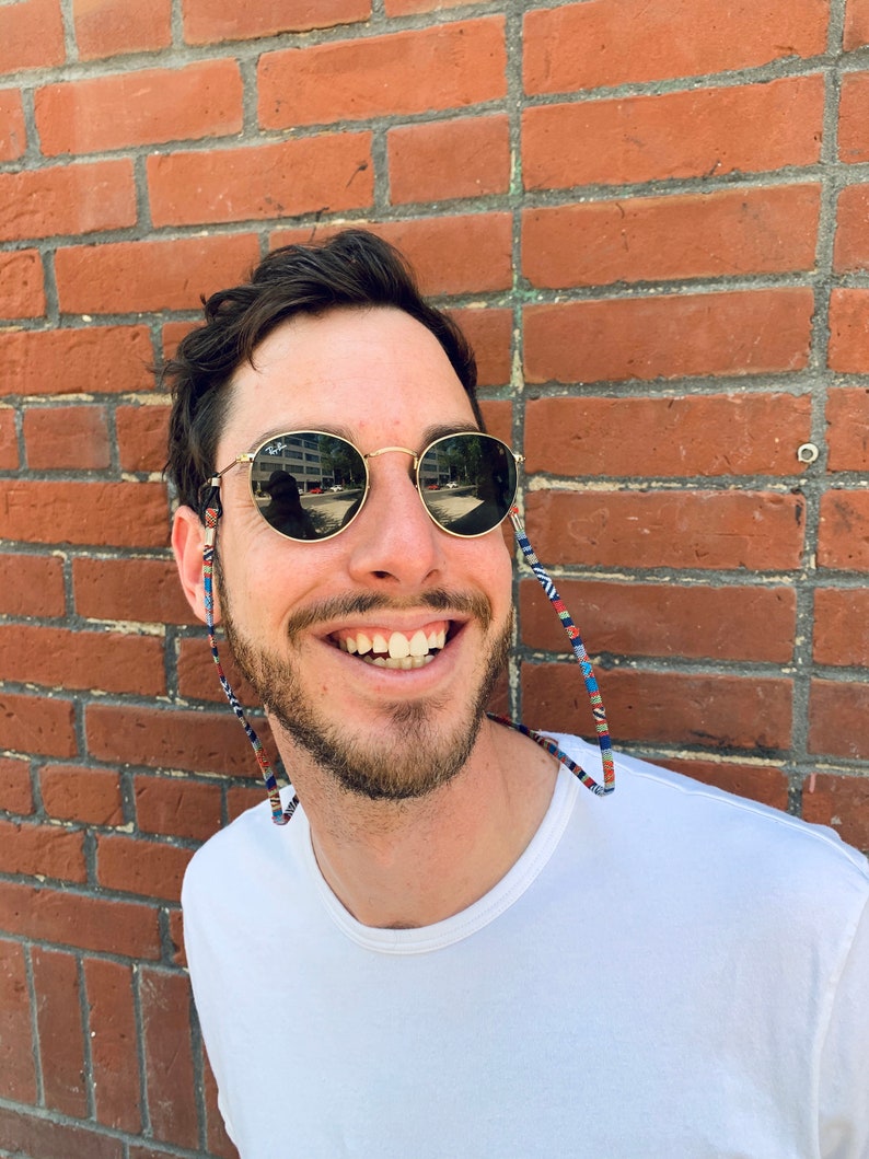 May include: A man with a beard is smiling and wearing round, dark sunglasses with a colorful patterned strap. He is wearing a white t-shirt and standing in front of a brick wall.