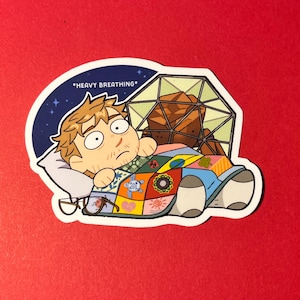 K&ouml;nnte beinhalten: Ein Aufkleber mit einer Zeichentrickfigur mit hellbraunem Haar, die unter einer bunten Patchwork-Decke liegt. Die Figur hat weit aufgerissene Augen und dar&uuml;ber steht der Text *HEAVY BREATHING*. Hinter der Figur befindet sich eine geometrische Kuppelstruktur.