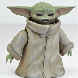May include: A green and brown plush toy of a character from the Star Wars franchise. The toy is wearing a brown coat with a fur collar and has large ears.
