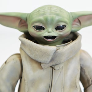 May include: A green and brown plush toy of a character from the Star Wars franchise. The toy has large ears, big eyes, and a toothy grin. The toy is wearing a brown, hooded robe.