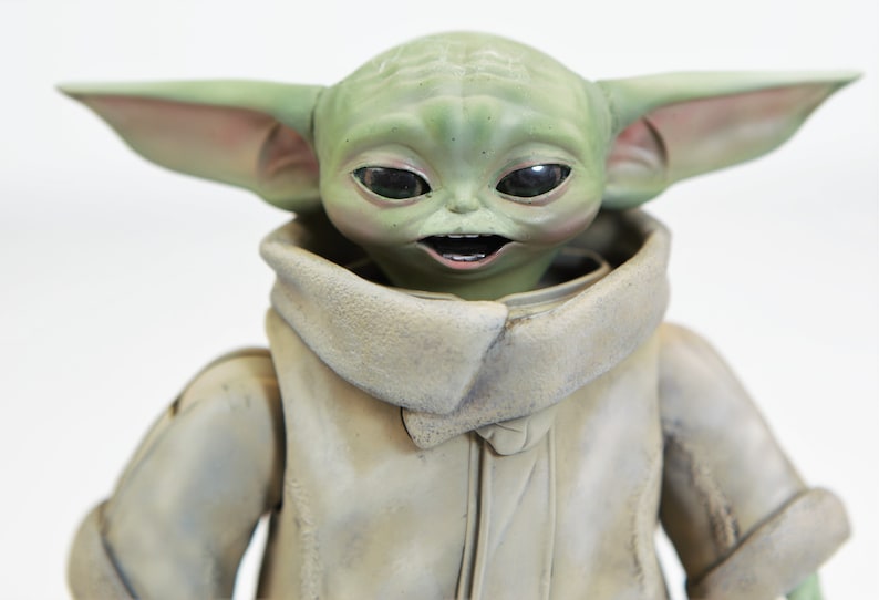 May include: A green and brown plush toy of a character from the Star Wars franchise. The toy has large ears, big eyes, and a toothy grin. The toy is wearing a brown, hooded robe.