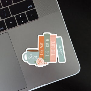 Puede incluir: Una pegatina de una pila de libros con las palabras "The Way", "The Truth", "The Life" y "John 14:6" en los lomos. Una taza de caf&eacute; con la palabra "Jesus" est&aacute; delante de los libros. Hay flores delante de los libros.