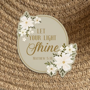 May include: A round sticker with a beige background and white flowers. The sticker has the text "Let your light shine" in gold lettering and "Matthew 5:16" in smaller gold lettering.