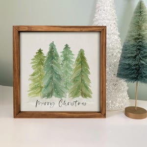 May include: A framed watercolor painting of green Christmas trees with white accents and the words "Merry Christmas" in a script font. The frame is made of brown wood. Decorative Christmas trees are in the background.