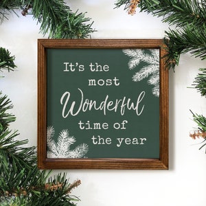 May include: A wooden sign with a green background and white text that reads "It's the most wonderful time of the year". The sign is decorated with white pine branches.