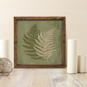 May include: A framed print featuring two fern fronds in shades of green and beige against a muted green background. The artwork is encased in a dark brown wooden frame, creating a natural and earthy aesthetic. The print is displayed on a wooden surface.