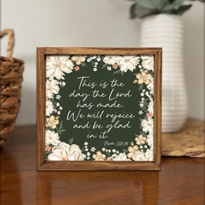 May include: A framed square print with a dark green background and white floral accents. The text reads, "This is the day the Lord has made. We will rejoice and be glad in it. Psalm 118:24." The frame is made of wood.