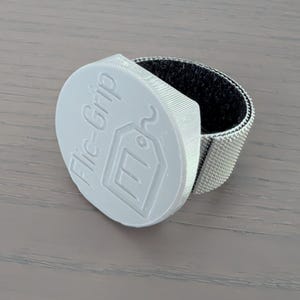 May include: A light grey Flic-Grip with a tag logo is attached to a silver and black adjustable strap. The product is designed to hold items and features the text "Flic-Grip".