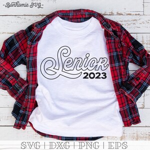 May include: White t-shirt with a black outline of the word "Senior" with the year "2023" underneath. The shirt is laid on a red and black plaid shirt.
