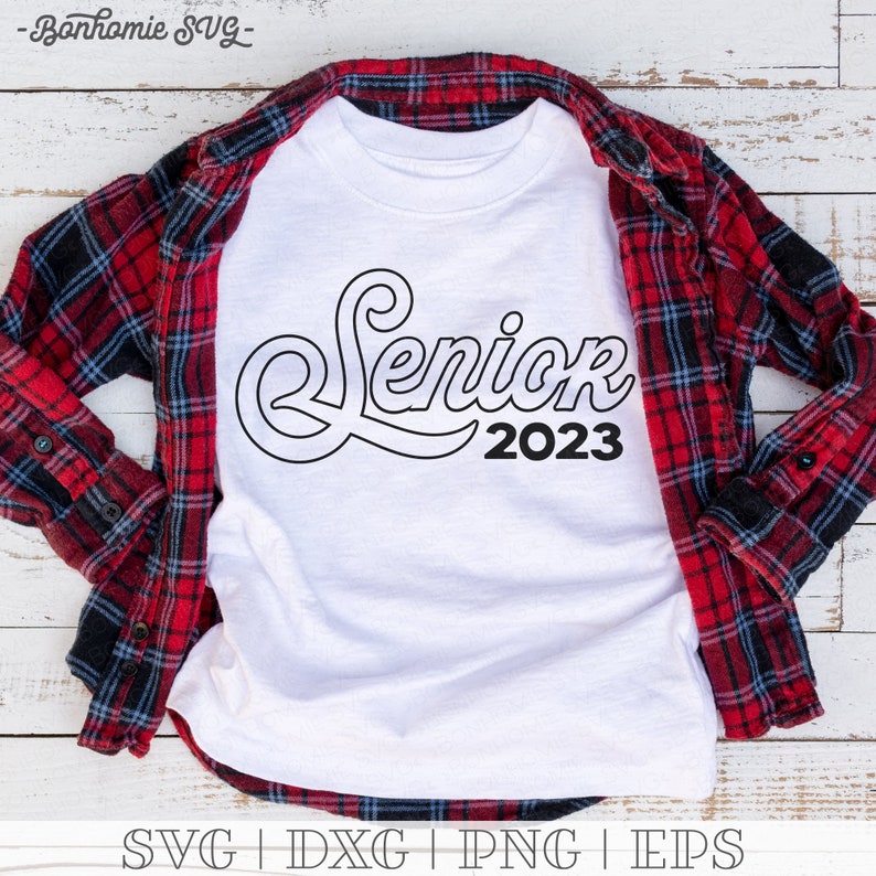 May include: White t-shirt with a black outline of the word "Senior" with the year "2023" underneath. The shirt is laid on a red and black plaid shirt.