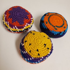 May include: Three round, hand-crocheted footbags featuring intricate, multi-colored geometric patterns. One displays purple, orange, and yellow; another has orange, blue, and black; and the third features yellow, purple, and blue with black speckles.