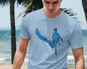 Może przedstawiać: Jasno niebieski t-shirt z grafiką przedstawiającą nurka w niebieskim kombinezonie i płetwach. Nurek trzyma harpun i jest otoczony bąbelkami. Koszulkę nosi osoba stojąca na plaży.