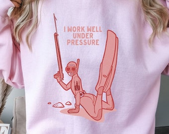 Könnte beinhalten: Rosafarbener Pullover mit einem Grafikdruck eines Tauchers und dem Text "Ich arbeite gut unter Druck".