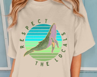 May include: A beige t-shirt with a green and blue graphic of a whale jumping out of the water. The graphic is surrounded by a circle with the text "Respect the Locals" in green.