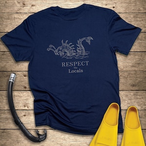 Könnte beinhalten: Marineblaues T-Shirt mit einem Seeungeheuer-Motiv und dem Text "Respect the Locals". Ein schwarzer Schnorchel und gelbe Flossen liegen auf einer Holzoberfläche.