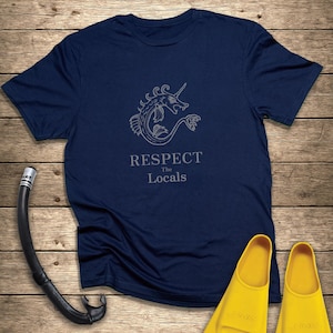 Può includere: Maglietta blu navy con grafica di creatura marina e la scritta "RESPECT The Locals". L'immagine include anche un boccaglio nero e pinne gialle, il tutto su uno sfondo di legno.