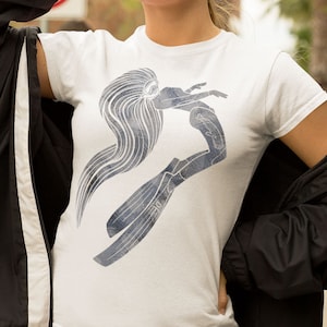 Puede incluir: Camiseta blanca con una ilustración estilizada de un buceador con pelo largo y aletas en tonos azules y grises. El gráfico está centrado en la parte delantera de la camiseta.