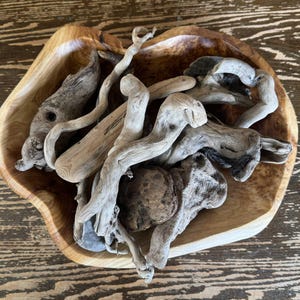 May include: A wooden bowl filled with various pieces of driftwood. The driftwood pieces are light gray and brown, with interesting shapes and textures. The bowl is made of light-colored wood and sits on a wooden surface.
