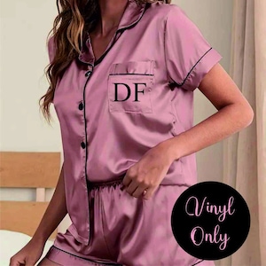 May include: A pink satin pajama set with short sleeves and shorts. The top has a black trim and a pocket with the initials "DF" embroidered on it. The shorts have a black trim and an elastic waistband. The text "Vinyl Only" is visible in the lower right corner of the image.