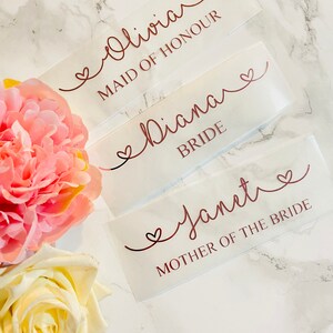 May include: Three white labels with rose gold lettering and hearts. The labels read "Olivia Maid of Honour", "Diana Bride", and "Janet Mother of the Bride".