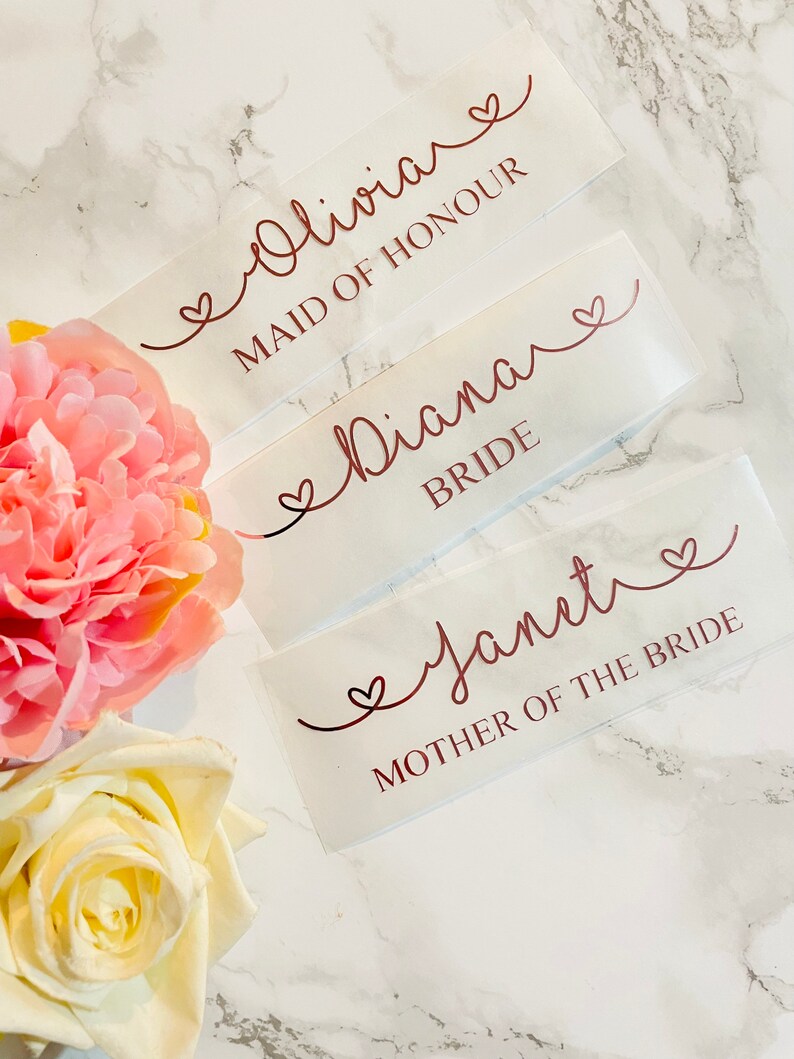 May include: Three white labels with rose gold lettering and hearts. The labels read "Olivia Maid of Honour", "Diana Bride", and "Janet Mother of the Bride".