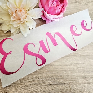 May include: A pink vinyl decal with the name "Esmee" in a cursive font. The decal is on a white backing, with a light wood grain background. Two flowers, one white and one pink, are in the background.