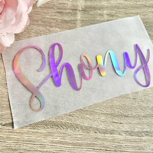 May include: A holographic vinyl decal with the name "Shony" in a cursive font. The letters transition through a spectrum of colors, including pink, yellow, and blue. The decal is on a translucent backing, with a wooden surface in the background.