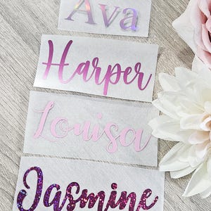 May include: Four name decals: "Ava" in iridescent, "Harper" in pink, "Louisa" in light pink, and "Jasmine" in glittery purple. Each name is in a cursive font, displayed on a white rectangular background. The decals are arranged on a light wood-grain surface.