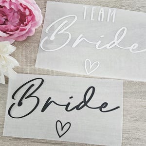 K&ouml;nnte beinhalten: Zwei durchscheinende Papierrechtecke mit den Worten "TEAM Bride" und "Bride" in Schreibschrift, jeweils mit einem Herzsymbol. Der obere Text ist wei&szlig;, der untere ist schwarz. Rosa und wei&szlig;e Blumen sind im Hintergrund.