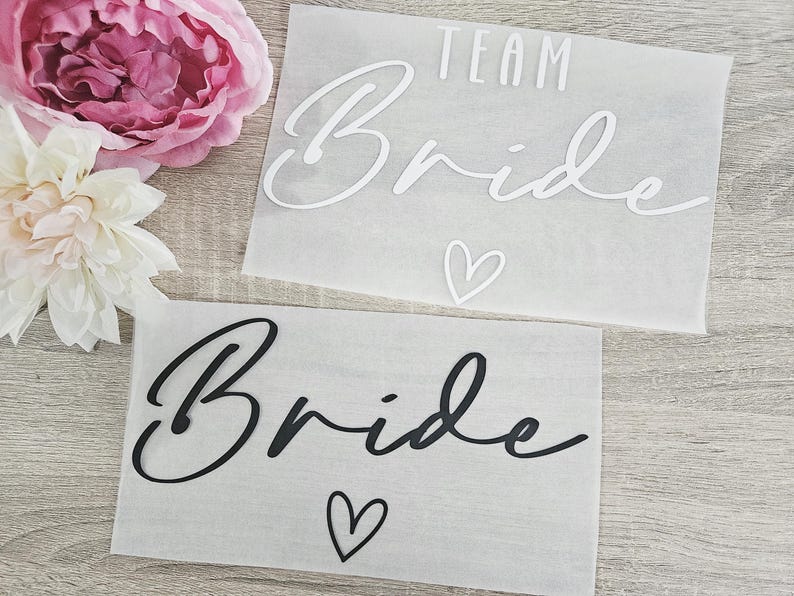K&ouml;nnte beinhalten: Zwei durchscheinende Papierrechtecke mit den Worten "TEAM Bride" und "Bride" in Schreibschrift, jeweils mit einem Herzsymbol. Der obere Text ist wei&szlig;, der untere ist schwarz. Rosa und wei&szlig;e Blumen sind im Hintergrund.