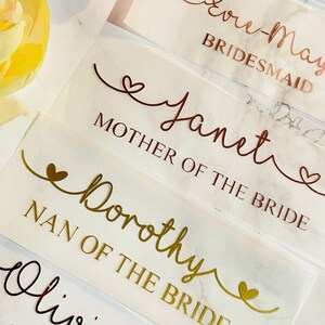 May include: White card stock place cards with gold and copper lettering. The cards have different titles for wedding party members, including "Bride's Maid", "Mother of the Bride", and "Nan of the Bride".