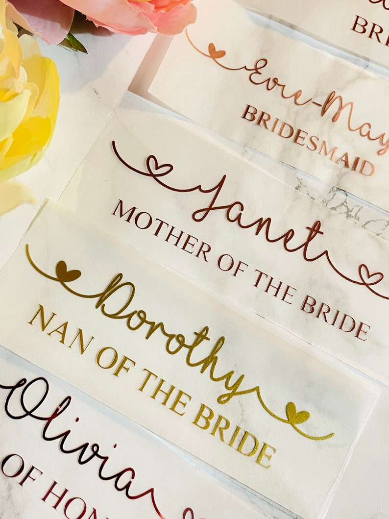 May include: White card stock place cards with gold and copper lettering. The cards have different titles for wedding party members, including "Bride's Maid", "Mother of the Bride", and "Nan of the Bride".