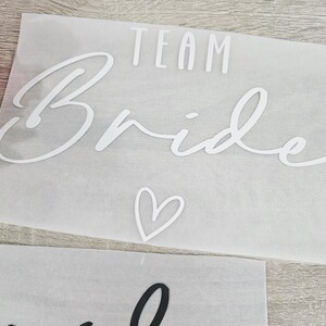 K&ouml;nnte beinhalten: Ein wei&szlig;er Aufkleber mit den Worten "TEAM Bride" in einer Schreibschrift, darunter ein Herzsymbol. Der Aufkleber befindet sich auf einem durchscheinenden Hintergrund. Eine rosa Blume ist in der oberen linken Ecke zu sehen.