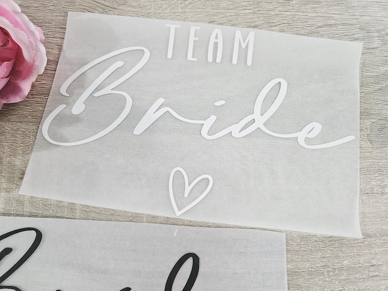 K&ouml;nnte beinhalten: Ein wei&szlig;er Aufkleber mit den Worten "TEAM Bride" in einer Schreibschrift, darunter ein Herzsymbol. Der Aufkleber befindet sich auf einem durchscheinenden Hintergrund. Eine rosa Blume ist in der oberen linken Ecke zu sehen.