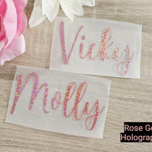 May include: Two personalized name decals, "Vicky" and "Molly", in rose gold holographic glitter script. The decals are on a white background, with a pink flower and a white flower in the background. The text "Rose Gold Holographic" is in a black rectangle.