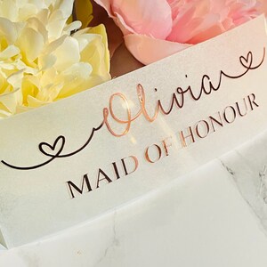 May include: A white card with the text "Olivia" in cursive with a heart symbol, and the text "Maid of Honour" in all caps.