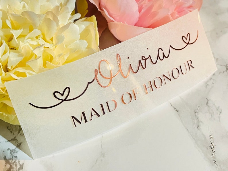 May include: A white card with the text "Olivia" in cursive with a heart symbol, and the text "Maid of Honour" in all caps.