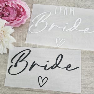 Puede incluir: Dos hojas de papel translúcido con las palabras "TEAM Bride" y "Bride" en escritura cursiva, acompañadas de símbolos de corazón. Una peonía rosa y una dalia blanca están en el fondo.