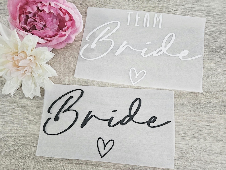 K&ouml;nnte beinhalten: Zwei durchscheinende Papierb&ouml;gen mit den Worten "TEAM Bride" und "Bride" in Schreibschrift, begleitet von Herzsymbolen. Eine rosa Pfingstrose und eine wei&szlig;e Dahlie befinden sich im Hintergrund.