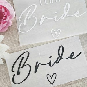 K&ouml;nnte beinhalten: Zwei rechteckige Bl&auml;tter mit dem Wort "Bride" in Schreibschrift und einem Herzsymbol. Ein Blatt hat das Wort "Team" &uuml;ber "Bride". Der Text ist auf einem Blatt wei&szlig; und auf dem anderen schwarz. Der Hintergrund ist durchscheinend wei&szlig;.