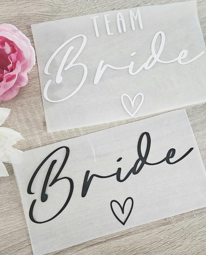 K&ouml;nnte beinhalten: Zwei rechteckige Bl&auml;tter mit dem Wort "Bride" in Schreibschrift und einem Herzsymbol. Ein Blatt hat das Wort "Team" &uuml;ber "Bride". Der Text ist auf einem Blatt wei&szlig; und auf dem anderen schwarz. Der Hintergrund ist durchscheinend wei&szlig;.