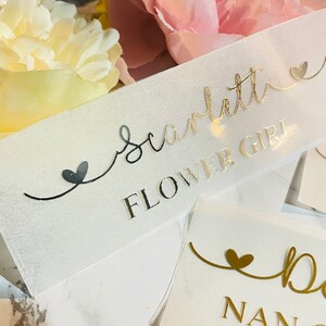 May include: A clear plastic sign with gold lettering that reads "Scarlett Flower Girl" with a small heart on either side of the text.