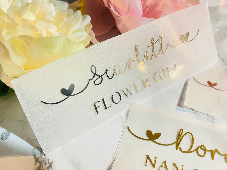 May include: A clear plastic sign with gold lettering that reads "Scarlett Flower Girl" with a small heart on either side of the text.
