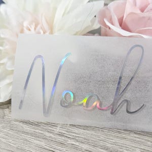 May include: A rectangular, transparent sticker with the name "Noah" in a holographic, cursive font. The sticker has a rainbow sheen and is set against a backdrop of white and pink flowers.