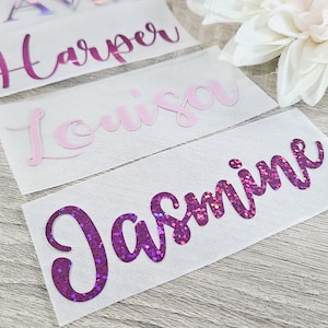 May include: Close-up of three name decals: "Harper" in magenta, "Louisa" in light pink, and "Jasmine" in glittery purple. The decals are on white backing, with a white flower in the background. The names are in a cursive font.
