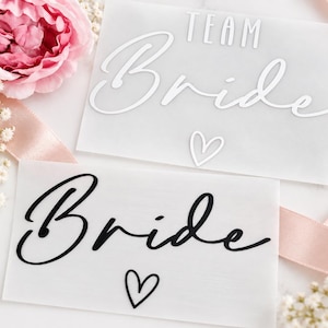 Op de afbeelding: Twee rechthoekige bordjes van mat acryl. Het bovenste bordje heeft witte tekst met TEAM Bride en een klein hartje. Het onderste bordje heeft zwarte tekst met Bride en een klein hartje. Beide bordjes zijn plat op een ondergrond geplaatst.
