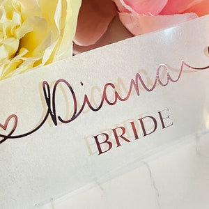 May include: A white satin ribbon with the name "Diana" written in rose gold script with a heart on either side. The words "Bride" are written in rose gold below the name.